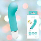 A turquoise VeDO 'Gee' Mini G-Spot Rechargeable Waterproof Vibrator with a curved tip for G-spot stimulation and a white base is displayed against a softly blurred background with pastel bokeh circles. To the right, there is the product's packaging, featuring an image of the vibrator and the text "VeDO gee MINI VIBE," boasting 10 modes of vibration.